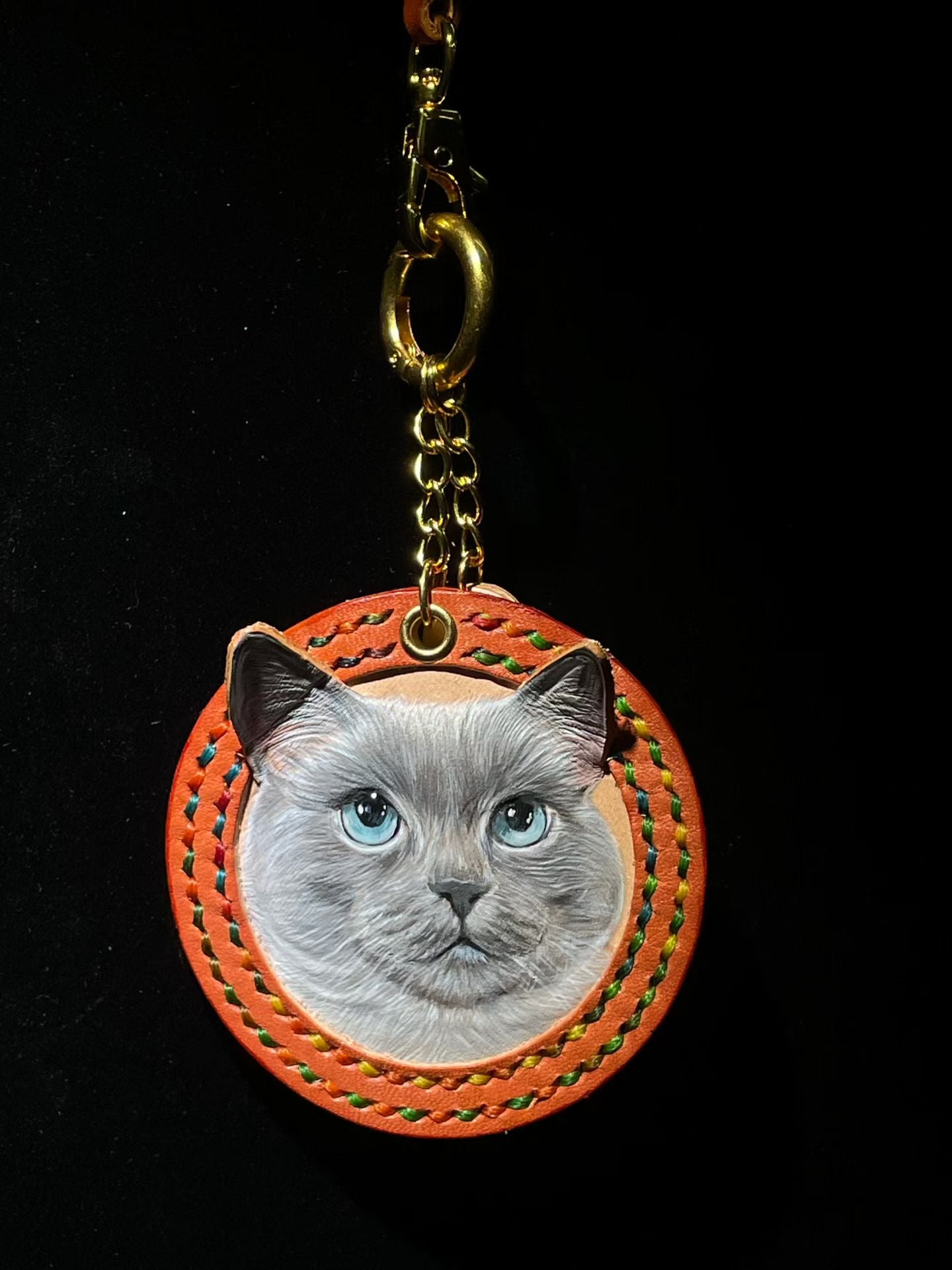 Supplier photograph of a grey cat portrait on an orange round charm with rainbow stitching.